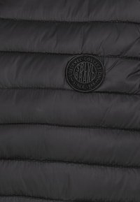 Schwarzer, gestepptes Material mit horizontalen Nähten und einem runden schwarzen Patch, auf dem steht "SBLVL Brand Sublevel Collection Since 1986".