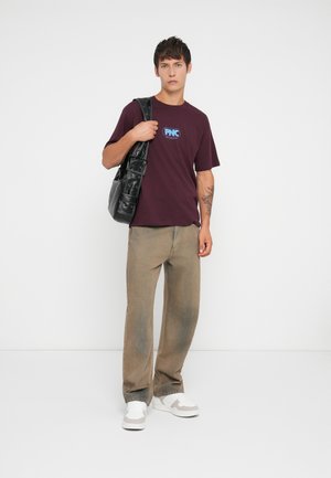 Maroon cotton t-shirt with a blue logo, paired with beige straight-leg pants. Black bag features multiple pockets, and white sneakers complete the look.