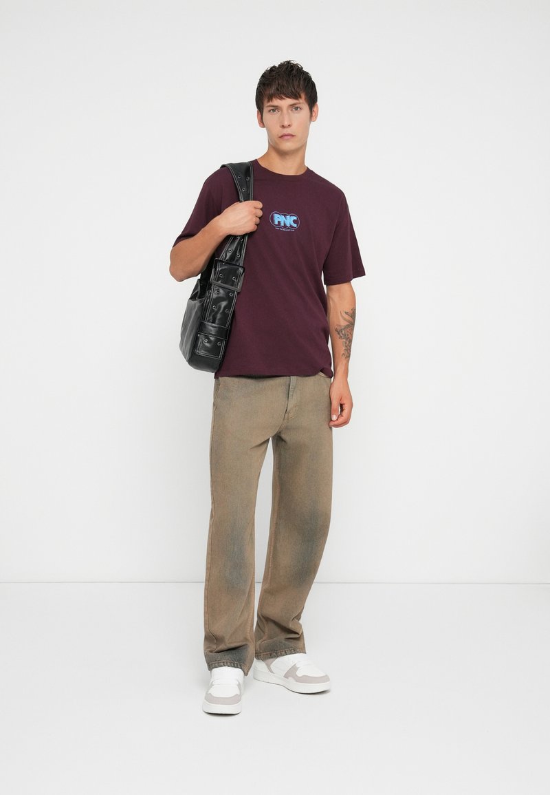 Maroon cotton t-shirt with a blue logo, paired with beige straight-leg pants. Black bag features multiple pockets, and white sneakers complete the look.