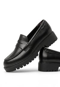 Ζευγάρι μαύρα δερμάτινα loafers με χοντρές σόλες με πέλμα και στρογγυλεμένες τετράγωνες μύτες, στοιβαγμένα με το ένα παπούτσι να ακουμπάει στη φτέρνα του άλλου.