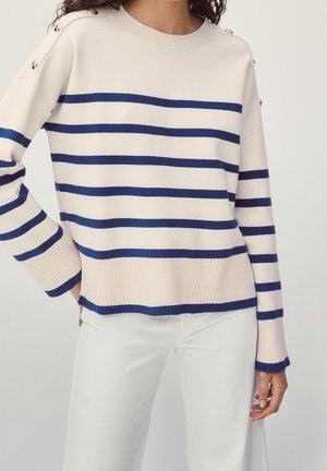 Woman wearing cream sweater with navy horizontal stripes and gold buttons on shoulders, paired with white pants, standing with hand on hip.