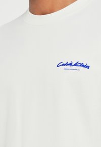 Balta medvilninė crewneck marškinėliai su mėlynu „Calvin Klein“ logotipu ant krūtinės. Audinys yra lygus ir minkštas.