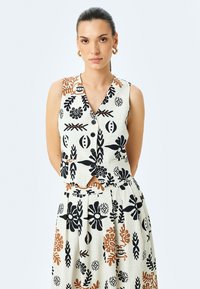 Top sin mangas color crema con botones, decorado con patrones florales en negro y óxido, combinado con una falda plisada a juego que presenta diseños similares.