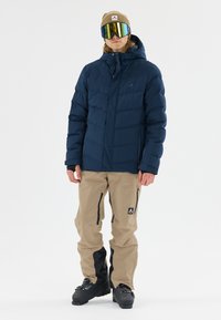 Doudoune bleue marine avec des coutures horizontales, capuche et poches zippées, associée à un pantalon softshell beige avec des accents et des zips noirs.