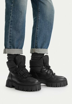 Beine in umgeschlagenen blauen Jeans, schwarzen, dicksohligen Schnürstiefeln und schwarzen Socken vor einem einfarbigen Hintergrund.