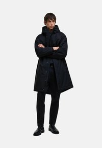 Fekete, vízálló parka kapucnival, elöl nyomógombos záródással, oldalsó zsebekkel és sima felülettel. Fekete nadrág és csizma fölött viselve.