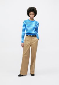 Pull tricot rayé bleu avec une texture côtelée, associé à un pantalon beige à jambes larges et une ceinture noire. Le mannequin se tient les mains dans les poches.