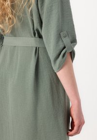 Vestido de tecido texturizado verde com cintura justa e mangas dobradas, apresentando um detalhe de botão na manga.