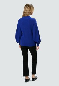 Blau gestrickter Pullover mit lockerer Passform und Ballonärmeln, kombiniert mit schwarzen Schlaghosen mit Spitzenbesatz am Saum.