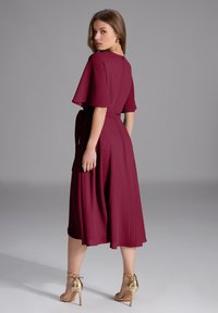 Robe midi bordeaux avec des manches fluides et une taille nouée. Texture lisse, silhouette en A, associée à des sandales à talons hauts dorées.