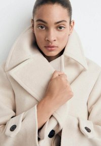 Jasno-beżowy wełniany płaszcz z szerokim kołnierzem, podwójnym zapięciem oraz dużymi czarnymi guzikami. Teksturowana tkanina o dopasowanym kroju.