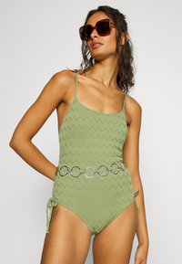 Maillot de bain une pièce vert avec une texture en zigzag, des bretelles ajustables et des liens sur le côté. Assorti d'une ceinture en cercle métallique et de lunettes de soleil surdimensionnées.
