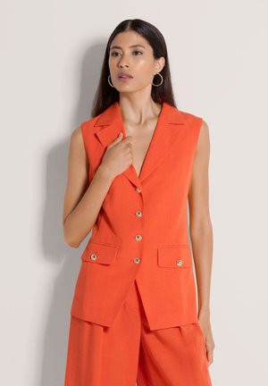 Vrouw met lang donker haar, draagt grote oorringen en een oranje mouwloos button-up blazer met bijpassende wijde broekspijpen.
