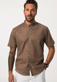 Camicia button-up a maniche corte in cotone marrone con colletto alla mandarino, caratterizzata da una trama testurizzata e bottoni in tonalità argento, abbinata a pantaloni bianchi.