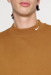 Detailní záběr na osobu s hnědým tričkem s kulatým výstřihem a malým bílým logem Nike ve tvaru klíče blízko límce.