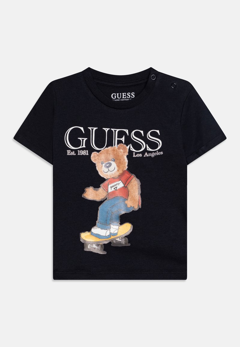 Musta orgaaninen puuvillapaita, jossa on kuva karhusta skeittilaudalla, valkoiset "GUESS"-tekstit ja punaisia yksityiskohtia karhun paidassa.