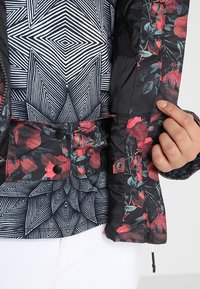 Schwarze Blumenjacke mit roten und grünen Mustern, Knopfverschlüssen und einem geometrisch gemusterten Innenfutter. Texturierter Stoff und detaillierte Akzente.