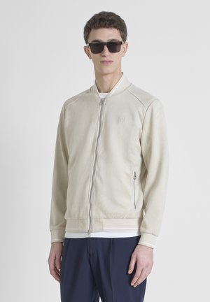 Chaqueta bomber de color beige claro con cremallera, hecha de un material suave, con puños acanalados, bolsillos laterales y un logo en relieve en el pecho.
