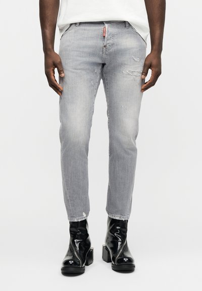 Homme portant un jean gris clair déchiré et des bottines à talons noires brillantes, debout les mains détendues le long du corps.