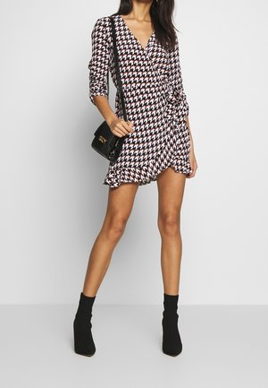 Femme portant une robe portefeuille pied-de-poule noir, blanc et rose avec un ourlet à volants, des bottines noires et tenant un sac à main noir à fermoir doré.
