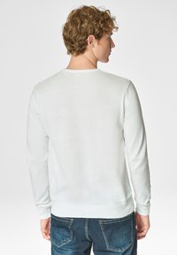 Maglione bianco a maniche lunghe con scollo rotondo, polsini e orlo a coste. Indossato con jeans in denim scuro, che mostrano una texture liscia e morbida.