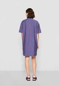 Vestido de manga curta em azul com padrão de riscas verticais em vermelho e roxo. Apresenta um colarinho alto e comprimento até à metade da coxa. Usado com ténis cor de vinho.