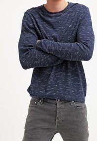 Chemise à manches longues bleu marine avec un motif texturé, col rond et coupe décontractée, associée à un jean gris. Aucun logo ni motif visible.