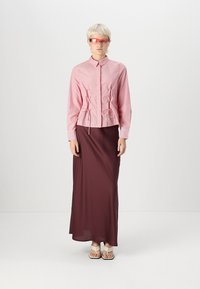 Črtasta rdeča in bela srajca s cinčnim pasom, v kombinaciji z burgundskimi maksi krilom. Model nosi rdeče sončna očala in lahke sandale.