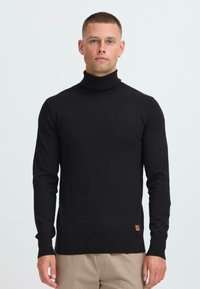 Modelo masculino con el pelo corto, vistiendo un suéter negro de cuello alto y pantalones beiges, de pie contra un fondo claro y liso.
