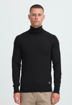 Modelo masculino con el pelo corto, vistiendo un suéter negro de cuello alto y pantalones beiges, de pie contra un fondo claro y liso.