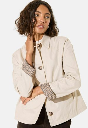 Vrouw met kort krullend haar draagt een beige jas met geruite manchetten, poseert met één hand bij de nek en de andere gekruist bij de taille.