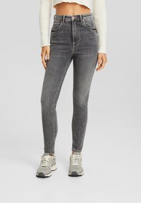 Jeans de mezclilla gris de cintura alta con un corte ajustado, costuras en contraste, bolsillos frontales y un lavado ligeramente desgastado. Combinados con zapatillas de color claro.