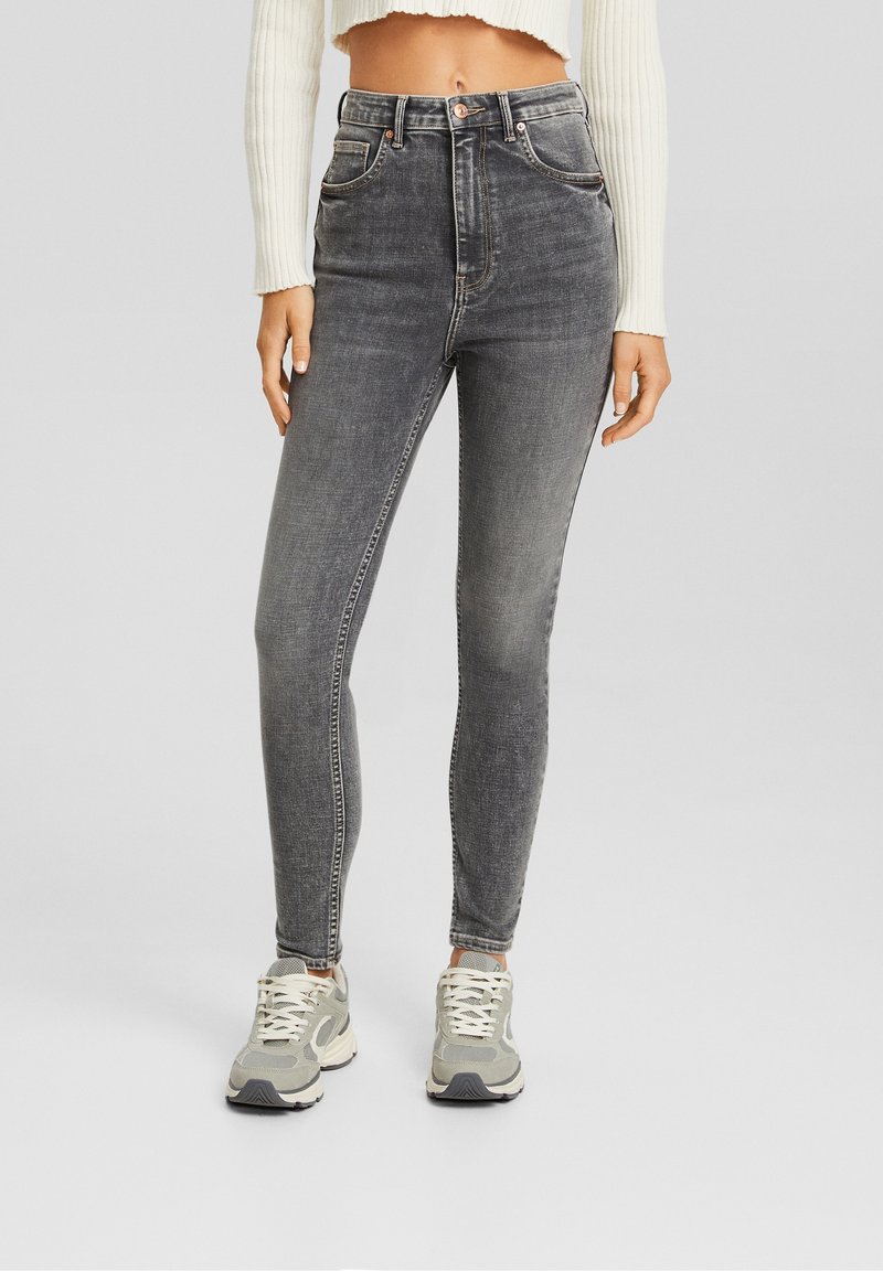 Jeans de mezclilla gris de cintura alta con un corte ajustado, costuras en contraste, bolsillos frontales y un lavado ligeramente desgastado. Combinados con zapatillas de color claro.