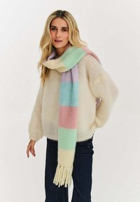 Kobieta ubrana w kremowy, puszysty sweter, ciemne spodnie i pastelowy szalik w paski z frędzlami, stojąca na tle jednolitego białego tła.