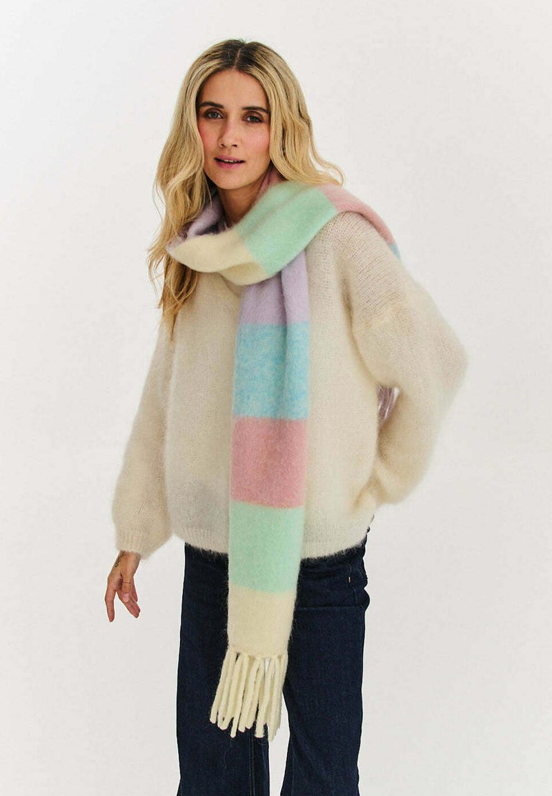 Kobieta ubrana w kremowy, puszysty sweter, ciemne spodnie i pastelowy szalik w paski z frędzlami, stojąca na tle jednolitego białego tła.