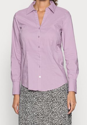 Mujer vistiendo una camisa lavanda de manga larga con botones y cuello, y una falda con patrón en blanco y negro.