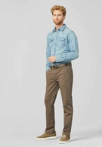 Lichtblauwe denimoverhemd met twee borstzakken, gecombineerd met een bruine broek. Beige suède sneakers met witte zolen, tegen een neutrale achtergrond.