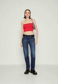 Ung kvinne iført rød stroppeløs crop top, blå jeans med svart belte, svarte platåsko og bærende en sølvfarget skulderveske på ensfarget bakgrunn.