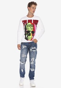 Langärmliges weißes Hemd mit buntem Schädel-Graphic, Text "1996 New York", kombiniert mit zerrissenen blauen Jeans und weißen Sneakers.