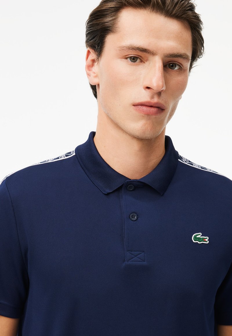 Marineblaues Poloshirt mit klassischem Kragen, zwei Knöpfen und strukturierter Optik. Verfügt über ein kleines grünes Krokodil-Logo auf der Brust.