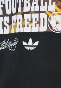 Μερική όψη μαύρου υφάσματος με λευκό κείμενο 'FOOTBALL IS FREED', σύμβολο χειραψίας, υπογραφή και λογότυπο Adidas.