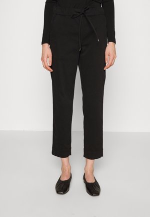 Mujer que lleva pantalones negros cortos con cintura de cordón y zapatos planos negros, de pie sobre un suelo gris claro.