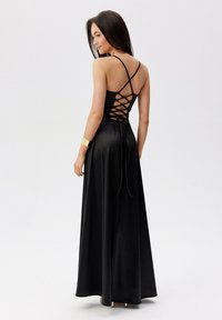 Černé saténové maxi šaty s přiléhavým vrškem, kříženými ramínky na zádech a detailem na vázání. Hladká textura s volným siluetem.