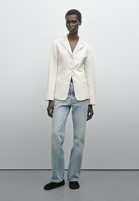 Hellbeige Blazer mit Reverskragen und fünf Knöpfen, kombiniert mit hellblauen, geraden Jeans und schwarzen Schuhen. Weiche Textur durchgehend.