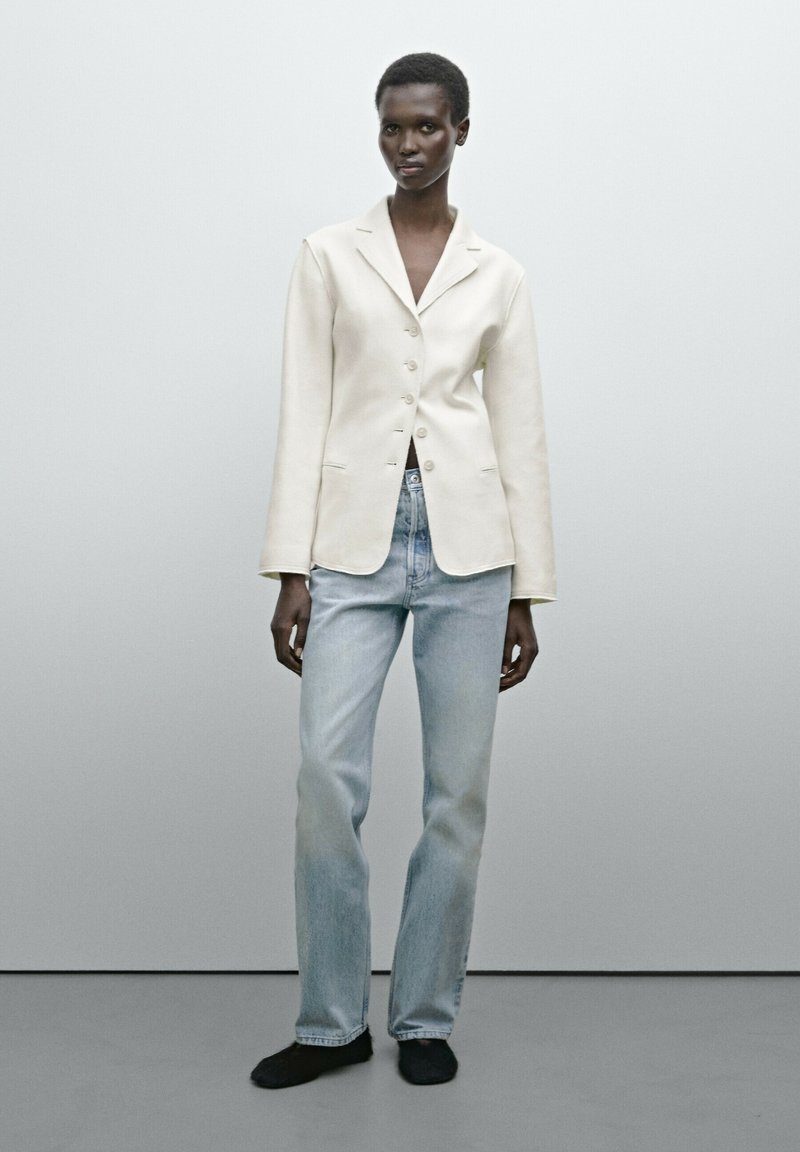 Blazer chiaro in crema con colletto a rever e cinque bottoni, abbinato a jeans dritti blu chiaro e scarpe nere. Texture morbida in tutto il capo.