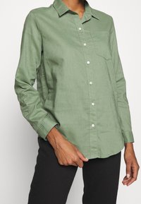 Chemise vert clair à manches longues, boutonnée avec col, poche poitrine et boutons blancs, fabriquée à partir d'un tissu doux et texturé.