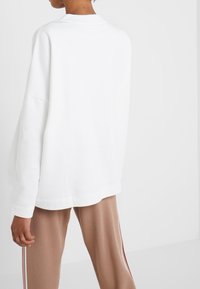 Sudadera blanca de gran tamaño con cuello redondo acanalado y hombros caídos, combinada con pantalones beige con rayas laterales. Textura de tela suave.