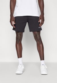 Black cotton shorts with a classic fit, featuring a white embroidered logo on the left leg, paired with white sneakers and ribbed socks.