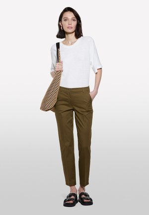 Weißes kurzärmliges T-Shirt, olivgrüne maßgeschneiderte Hose, gemusterte Tragetasche, schwarze Sandalen. Model steht mit einer Hand in der Tasche.