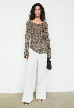 Frau mit lockigem Haar, die einen strukturierten braun-beigen Pullover, weiße Hosen mit weitem Bein, schwarze Sandalen trägt und eine schwarze geflochtene Handtasche hält.